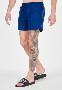 Pantaloni da bagno blu navy con vita elastica, cordoncino nero e strisce laterali verdi; indossati con sandali neri con logo.