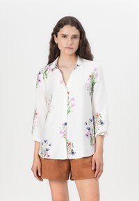 White button-up shirt with 3/4 sleeves, featuring colorful floral print on shoulders and sleeves, paired with brown suede shorts.
