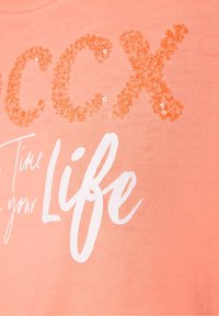 Camiseta de color durazno con letras en lentejuelas naranjas brillantes que dicen "CCX" y texto blanco que dice "Time your Life" en un estilo de escritura a mano.