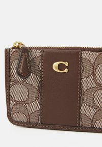 Portefeuille en cuir marron avec une toile à motifs, quincaillerie dorée, fermeture à zip et un détail de logo saillant à l'avant. Panneaux d'accent texturés.
