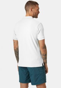 Polo bianco a maniche corte con tessuto texturizzato, fessure laterali e colletto. Indossato con pantaloni corti teal, che mostrano un look estivo casual.