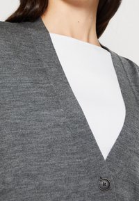 Szary kardigan z dekoltem w serek, wykonany z fakturowanej dzianiny. Posiada czarny guzik z przodu, noszony na białej bluzce.