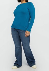 Turkosblå tröja med rund halsringning och långa ärmar, med ribbat nederkant, kombinerad med mörkblå utsvängda jeans; modellen bär krämfärgade sneakers.