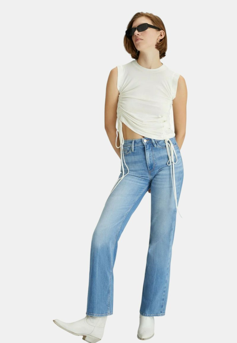 Top bianco senza maniche con arricciature laterali e lacci, abbinato a jeans a gamba dritta blu chiaro e stivaletti bianchi alla caviglia. Inclusi occhiali da sole neri.