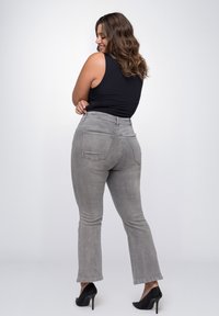 Grijze flared jeans met een hoge taille, voor- en achterzakken, gemaakt van denim, gecombineerd met zwarte hoge hakken. Model weergegeven van achteren.
