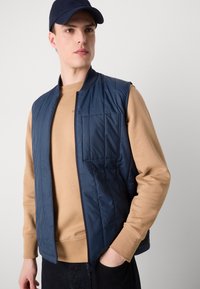 Colete acolchoado azul-marinho com fecho éclair na parte da frente, colarinho canelado e um bolso no peito, usado sobre um sweatshirt bege, contra um fundo simples.
