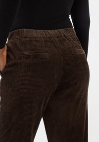 Brązowe spodnie z korduroy z teksturowanym, prążkowanym materiałem. Elastyczny pas, tylne kieszenie z haftowanymi akcentami. Zbliżenie widok z boku.