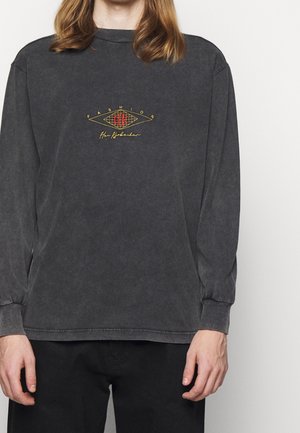 Person, der bærer en mørkegrå langærmet skjorte med gul og rød grafik med teksten "FASHION" og stiliseret tekst på brystet, kombineret med sorte bukser.