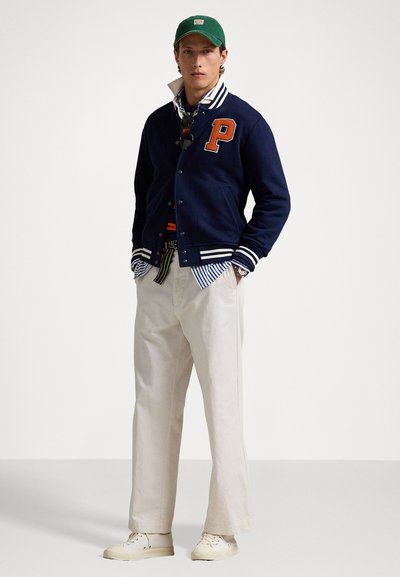 Námornícka univerzitná bunda s bielymi a oranžovými akcentmi, svetlo modrá a biela pruhovaná košeľa pod ňou, krémové nohavice a zelená čiapka. Biely sneakers.