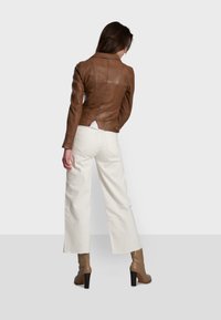Braune Lederjacke mit taillierter Passform, ausgestattet mit einem Reverskragen und sichtbaren Nähten, kombiniert mit weißen weiten Hosen und hohen Stiefeln.