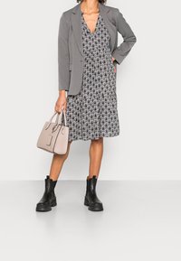 Blazer gris porté sur une robe noire et blanche à motifs, associée à des bottines noires à bouts ronds et un sac à main rose pâle avec des anses supérieures.