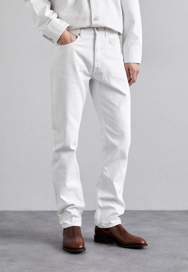Jean blanc en denim avec une coupe droite et des poches latérales, associés à des bottes en cuir brun à la cheville. Le tissu a une texture lisse.