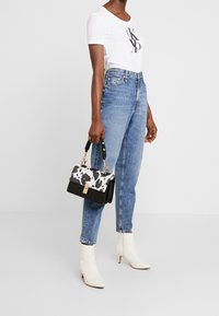 Kvinna i vit t-shirt med logotyp, högmidjade blå jeans, vita ankelskor, håller en svart och vit mönstrad handväska.