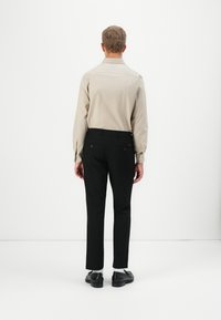Mann steht mit dem Rücken zur Kamera, trägt ein beigefarbenes Langarmhemd, schwarze Hosen, weiße Socken und schwarze Dressschuhe vor einem schlichten weißen Hintergrund.