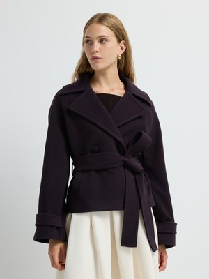 Lila Doppelreiherjacke mit breiten Revers, einem Bindegürtel und weit ausgeformten Ärmeln, aus einem weichen Textil gefertigt. Kombiniert mit einem weißen Rock.