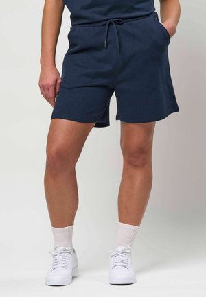 Isik, kes kannab tumesiniseid paelaga lühikesi pükse, heledaid roosakaid sokke ja valgeid tossusid ning seisab ühe käega taskus.