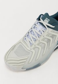Pantofii de tenis ușori, de culoare albă și turcoaz, cu o parte superioară din plasă, suprapunerile structurate, șireturi plate și o talpă texturată pentru aderență.