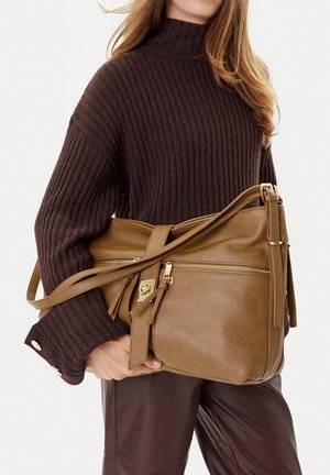 Mujer que lleva un suéter acanalado marrón oscuro y pantalones de cuero sostiene un gran bolso de cuero color beige con detalles de cremallera y hebilla dorados.