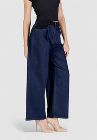 Jeans a palazzo blu scuro con vita alta, dotati di ampie tasche anteriori e una texture liscia, abbinati a tacchi alti beige.