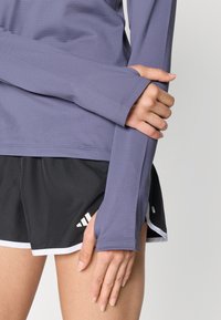 Haut de sport à manches longues de couleur violette avec une finition texturée et des trous pour les pouces, associé à un short noir avec des bordures blanches et des accents de logo.