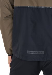 Wasserabweisende Jacke mit einem braunen oberen Teil und einem marineblauen unteren Teil, ausgestattet mit reflektierenden Akzenten und einem "ENDURANCE"-Label am Saum.