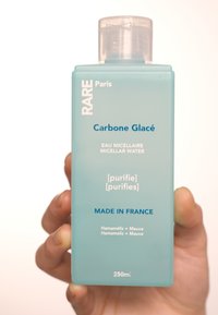 Bottiglia di plastica rettangolare trasparente contiene acqua micellare blu, etichettata "Carbone Glacé," con testo che indica gli ingredienti e il volume.
