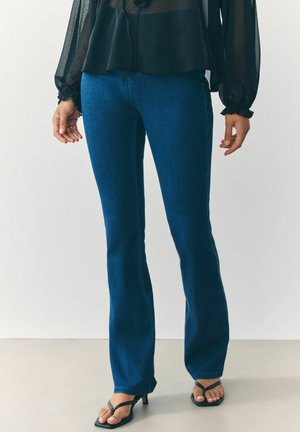 Blauwe flared jeans van denim, met een gemiddelde wasbeurt. Gecombineerd met een doorschijnende zwarte blouse en zwarte sandaaltjes met bandjes.
