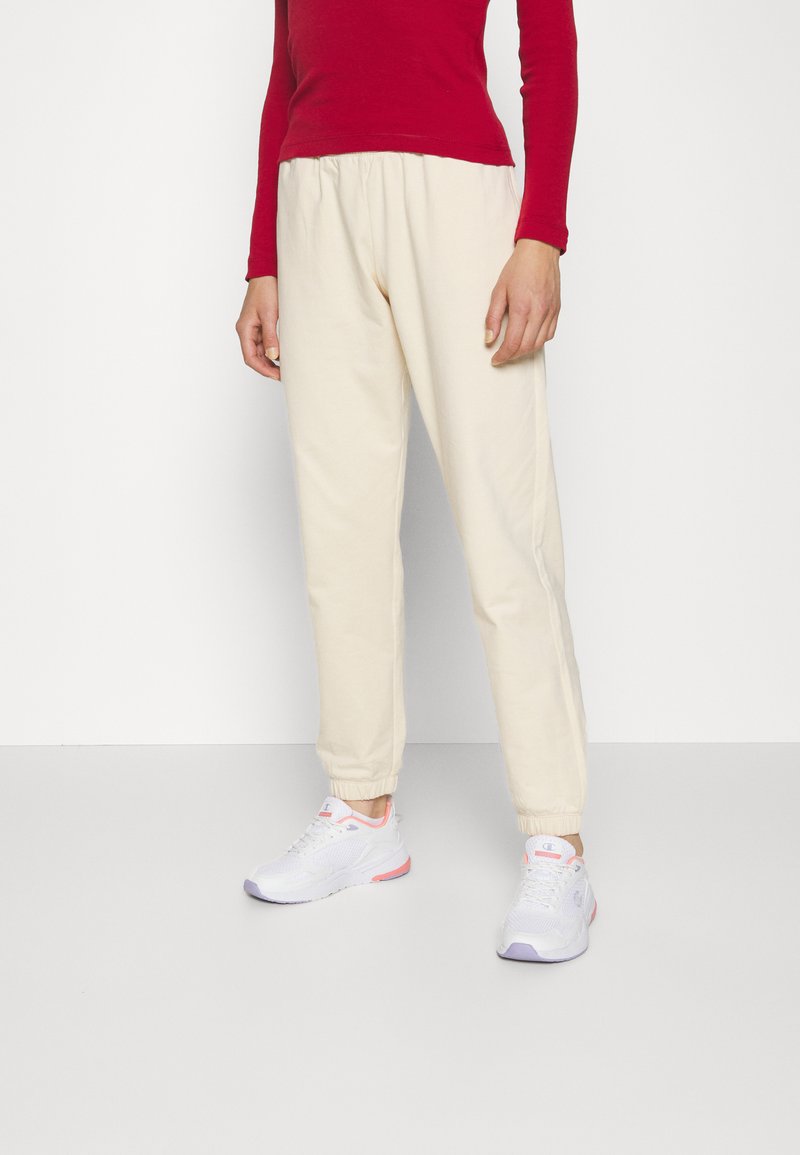 Pantalones de chándal color crema, ajustados en la parte inferior con cintura elástica y puños, combinados con zapatillas deportivas blancas que tienen acentos en rosa y morado.