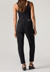 Femme portant un haut noir sans manches, un jean noir taille haute et des sandales à talons noires, debout de dos sur un fond uni.