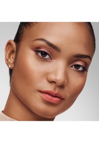 Lähetä lähikuva kasvoista, joilla on sileä, ruskea iho, pehmeästi muotoillut posket, vaaleanpunainen luomiväri ja mattapintainen korallihuuli. Yksinkertainen nappikorvakoru korostaa ilmettä.