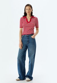 Polo shirt à rayures rouges et blanches à manches courtes avec un col en V, associé à un jean en denim bleu ample et des baskets beiges.