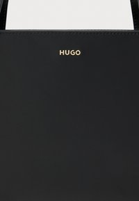 Sac à main synthétique noir à texture lisse, orné d'un logo "HUGO" doré. Comprend des poignées robustes et minimalistes pour un transport facile.