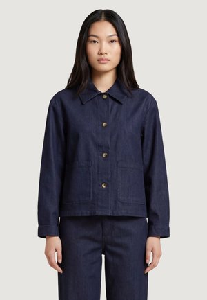 Femme aux longs cheveux noirs portant une veste boutonnée bleu foncé avec des poches à l'avant, debout devant un fond clair uni.