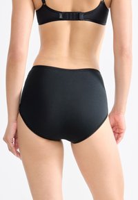Donna che indossa delle mutande nere a vita alta e un reggiseno nero abbinato, vista di spalle su uno sfondo bianco semplice.