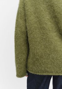 Oliwkowy, puszysty sweter o luźnym kroju i teksturowanym dzianiu. Posiada szerokie rękawy i okrągły dół, w połączeniu z ciemnoniebieskimi dżinsami.
