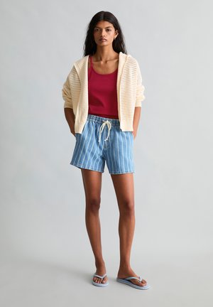 Giovane donna che indossa una felpa con cappuccio in maglia colore crema, canottiera rossa, pantaloncini a righe blu e infradito azzurre, in piedi davanti a uno sfondo grigio uniforme.