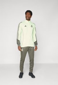 Light green athletic tracksuit with gray accents. Features zip-up jacket with black logo and matching tapered pants, made of smooth fabric.