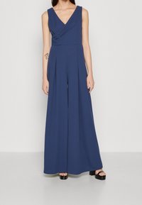 Marineblauer Jumpsuit mit V-Ausschnitt, tailliertem Oberteil und weit geschnittenen Hosenbeinen. Aus einem glatten Stoff mit Plissee-Details gefertigt. Schwarze Plateauschuhe.