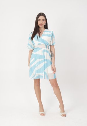 Frau mit langen dunklen Haaren, die ein blau-weiß gemustertes Wickelkleid mit kurzen Ärmeln und weiße Sandalen trägt, mit einem Bein nach vorne stehend.