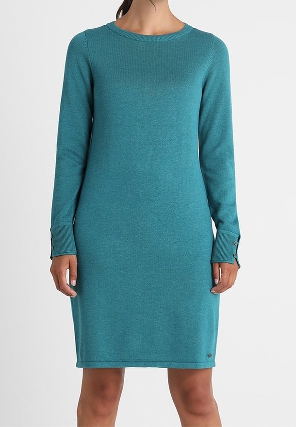 Femme portant une robe en maille bleu sarcelle à manches longues et longueur genou avec des détails de boutons aux poignets, sur un fond uni.