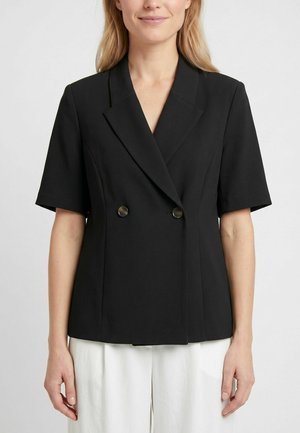 Mujer con blazer negro de manga corta y doble botonadura con solapas y pantalones blancos sobre un fondo claro y liso.