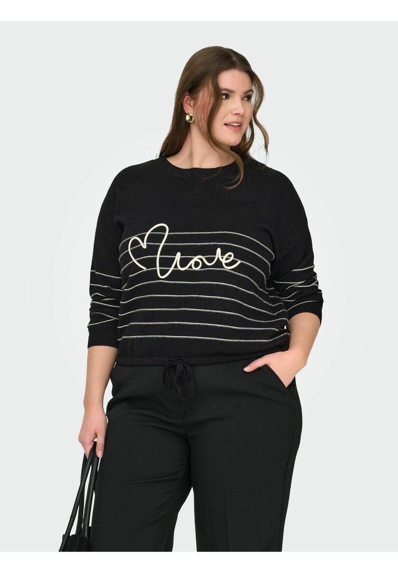 Femme portant un pull noir à rayures blanches avec le mot "love", tenant un sac à main noir, debout avec une main dans la poche.