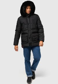 Jachetă puffer neagră cu glugă căptușită cu blană, având buzunare tip patch, închidere cu fermoar și un design matlasat, purtată cu blugi și cizme.