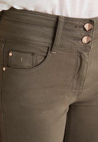 Jeans de mezcla de algodón verde oliva con cintura alta, que presentan dos botones en tono cobre, bolsillo frontal y detalles de costura sutil.