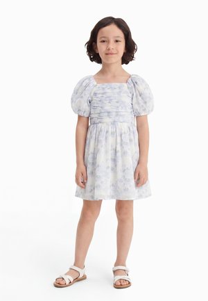 Jong meisje staat, draagt een witte jurk met opgeblazen mouwen en witte sandalen, glimlacht lichtjes tegen een eenvoudige witte achtergrond.