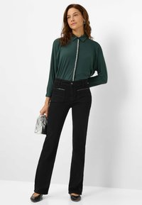 Maglia a maniche lunghe verde scuro con dettagli di bottoni lucidi, abbinata a pantaloni neri a vita alta con tasche anteriori. Il modello tiene una borsa argento.