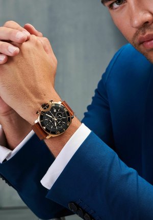 Montre chronographe ronde avec cadran noir, accents dorés et bracelet en cuir marron, portée sur un poignet avec un costume bleu et une chemise à manches blanches.