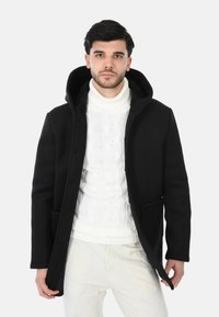 Cappotto nero con cappuccio dalla texture liscia, dotato di chiusura frontale con bottoni e tasche laterali, indossato sopra un maglione bianco a trecce.