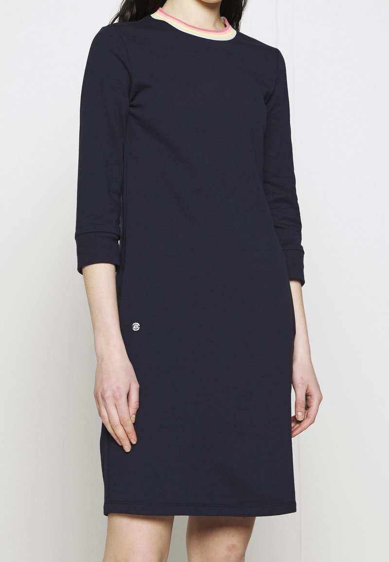 Femme portant une robe bleue marine au genou avec des manches trois quarts et un col rayé multicolore sur un fond uni.