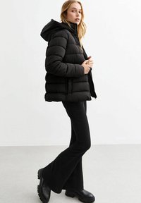 Veste en duvet noire avec capuche, dotée de sections matelassées et d'une fermeture éclair à l'avant. Association avec un pantalon évasé noir et des bottes noires robustes.